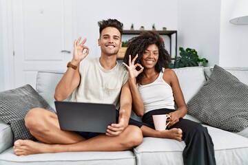 Young interracial couple using laptop at home sitting on the sofa smiling positive doing ok sign...