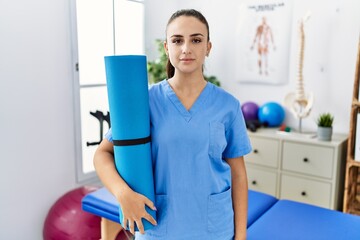 Young physiotherapist woman holding yoga mat thinking attitude and sober expression looking self confident