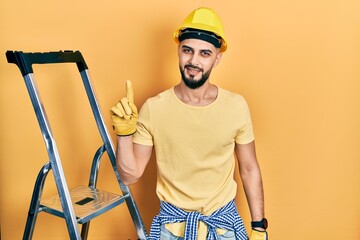 Handsome man with beard by construction stairs wearing hardhat pointing finger up with successful idea. exited and happy. number one.