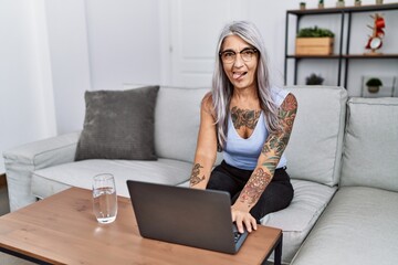 Middle age grey-haired woman using laptop at home sticking tongue out happy with funny expression....