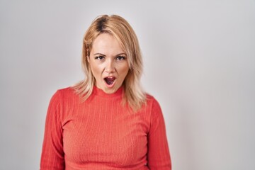 Blonde woman standing over isolated background in shock face, looking skeptical and sarcastic, surprised with open mouth