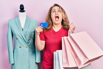 Middle age caucasian woman holding shopping bags and credit card angry and mad screaming frustrated and furious, shouting with anger looking up.