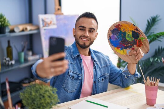 Young hispanic man artist make selfie by smartphone holding palette at art studio