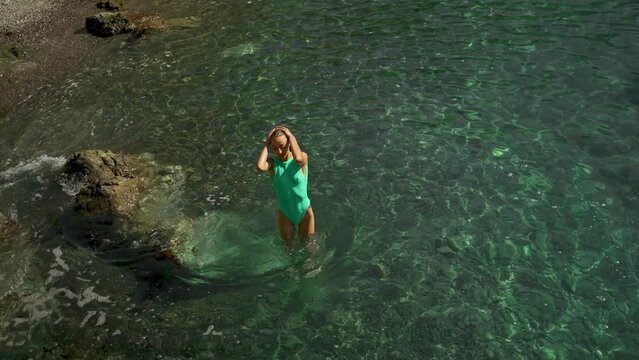 stunning slim fit body woman in green swimsuit relaxing in clear turquoise water of mediterranean sea after swimming. woman gets up from water and wipes drops from her face and hair.