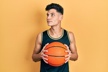 Young hispanic man holding basketball ball smiling looking to the side and staring away thinking.