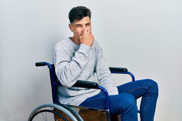 Fototapeta na wymiar Young hispanic man sitting on wheelchair smelling something stinky and disgusting, intolerable smell, holding breath with fingers on nose. bad smell
