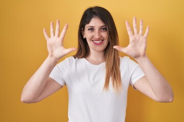 Young brunette woman standing over yellow background showing and pointing up with fingers number ten while smiling confident and happy.