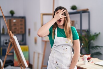 Young brunette woman at art studio peeking in shock covering face and eyes with hand, looking...