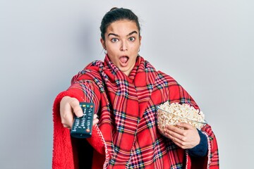 Young caucasian girl wearing blanket eating popcorn using tv control afraid and shocked with surprise and amazed expression, fear and excited face.