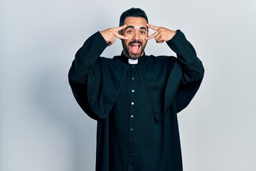 Handsome hispanic man with beard wearing catholic priest robe doing peace symbol with fingers over face, smiling cheerful showing victory