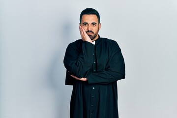 Handsome hispanic man with beard wearing catholic priest robe thinking looking tired and bored with depression problems with crossed arms.