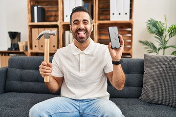 Young hispanic man with beard holding hammer and broken smartphone showing cracked screen sticking tongue out happy with funny expression.