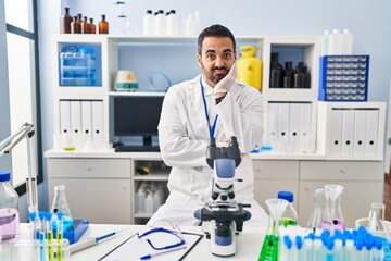 Young hispanic man with beard working at scientist laboratory thinking looking tired and bored with depression problems with crossed arms.