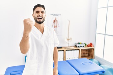 Young handsome man with beard working at pain recovery clinic angry and mad raising fist frustrated and furious while shouting with anger. rage and aggressive concept.