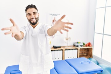 Fototapeta na wymiar Young handsome man with beard working at pain recovery clinic looking at the camera smiling with open arms for hug. cheerful expression embracing happiness.