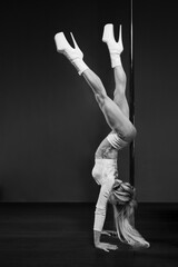 A muscular female dancer with long blonde hair is doing a Handstand Back Stretch move on a vertical pole in a dance studio. Pole dance workout.