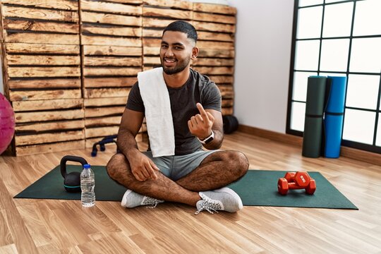 Young indian man sitting on training mat at the gym beckoning come here gesture with hand inviting welcoming happy and smiling