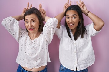 Hispanic mother and daughter together posing funny and crazy with fingers on head as bunny ears,...