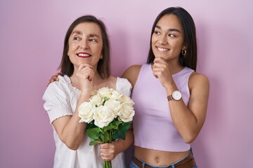 Hispanic mother and daughter holding bouquet of white flowers with hand on chin thinking about question, pensive expression. smiling and thoughtful face. doubt concept.