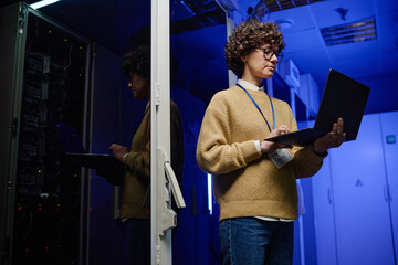 Young female engineer concentrating on her work of examining data on laptop standing in server room