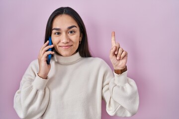 Young south asian woman having conversation talking on the smartphone smiling with an idea or question pointing finger with happy face, number one