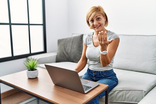Middle age blonde woman using laptop at home beckoning come here gesture with hand inviting welcoming happy and smiling