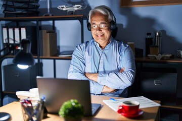 Hispanic senior man wearing call center agent headset at night happy face smiling with crossed arms looking at the camera. positive person.