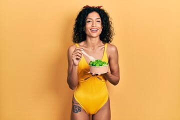 Naklejka premium Young latin girl wearing swimsuit eating salad smiling and laughing hard out loud because funny crazy joke.