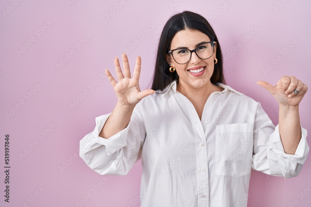 Poster Young brunette woman standing over pink background showing and pointing up with fingers number six while smiling confident and happy.