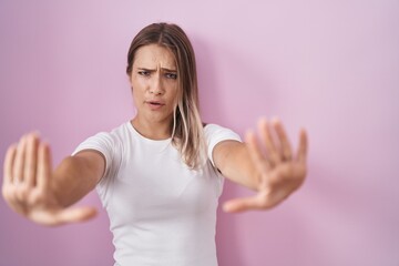 Blonde caucasian woman standing over pink background doing stop gesture with hands palms, angry and frustration expression