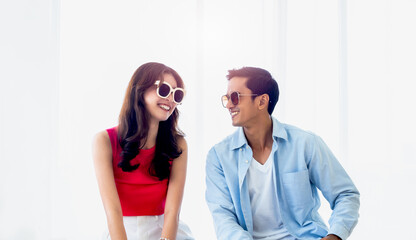 Happy Asian couple in summer, young man and woman wearing sunglasses with smile, sitting on the bed in the bright bedroom near the glass window, holiday travel, summer vacation concept.