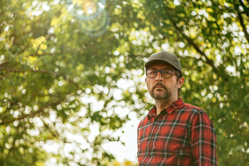 Portrait of male farmer wearing plaid shirt and trucker's hat posing in walnut orchard and looking...