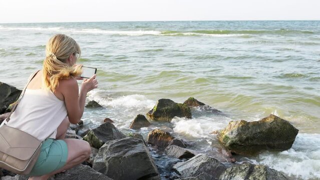 Woman photographs the sea landscape on the phone while sitting on the stones