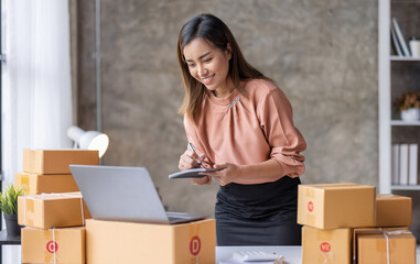 SME Online seller Young Asian woman working on laptop and box checking online order, check goods stock delivery package shipping postal. Asian woman startup SME small business at home office