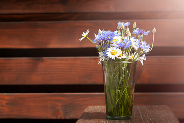 A bouquet of daisies and cornflowers in a glass vase on a wooden table against the background of a wooden wall. Place for an inscription. Selective focus. Copyspace.