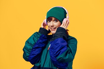 a happy, joyful woman in a green jacket and hat enjoys music standing in pink headphones on a yellow background holding them with her hands and looking at the camera