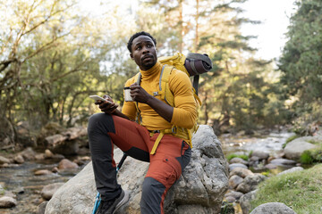 handsome young african american man with backpack smiling at sunset sitting on the rock, while...