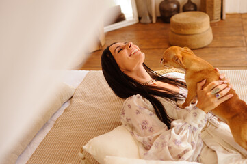 Happy stylish woman playing with Chihuahua dog on bed in bungalow at resort
