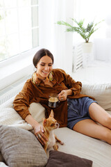 Positive stylish woman holding coffee cup and petting blurred Chihuahua dog on couch in living room 