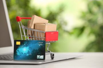 Online payment concept. Small shopping cart with bank card, boxes and laptop on white table, space...