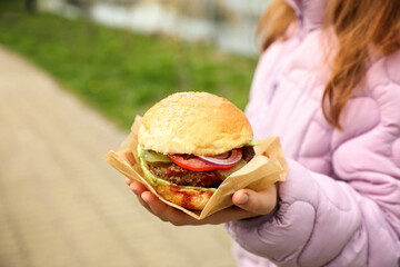 Little girl holding fresh delicious burger outdoors, closeup. Street food