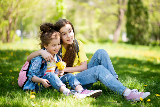 Two cute sisters' girls on a walk. It's spring outside and the sun is bright. A girl holds a dandelion in her hands. She is wearing a colored T-shirt and a denim shirt. Childhood. Family.