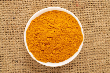 Aromatic seasoning turmeric in a white ceramic dish on burlap, close-up, top view.