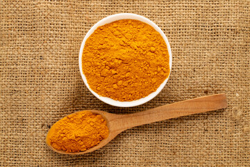 Aromatic seasoning turmeric in a white ceramic dish with a wooden spoon on burlap, close-up, top view.