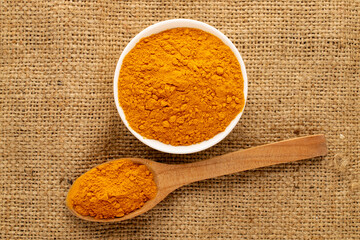 Aromatic seasoning turmeric in a white ceramic dish with a wooden spoon on burlap, close-up, top...