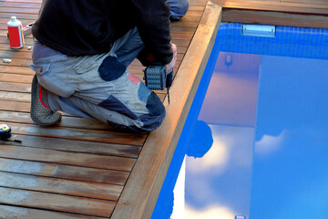 Man wearing working clothes is making a hole on the swimming pool wooden brown deck with the drilling machine. Aside is visible the blue clear water.