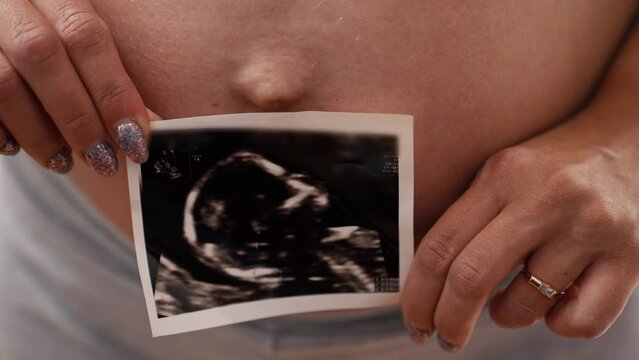 Nude big pregnant belly of a caucasian tattooed woman in gray biker shorts holding an ultrasound photograph of her unborn child. High quality 4k footage