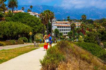 a couple walking wearing colorful clothes standing on a sidewalk surrounded by lush green trees, grass and plants and apartments and homes with blue sky and clouds at Leadbetter Beach in Santa Barbara