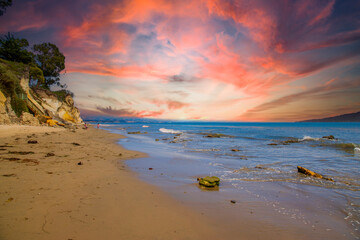 a gorgeous summer landscape at the beach with blue ocean water with waves rolling into the beach, silky brown sand and rocky cliffs covered in lush green trees and plants and powerful clouds at sunset