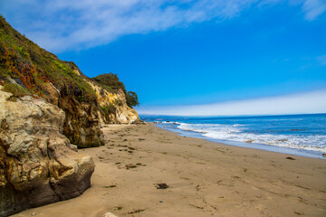 a gorgeous summer landscape at the beach with brown cliffs covered in lush green trees and plants and deep blue ocean water, blue sky and clouds at Leadbetter beach in Santa Barbara California USA - obrazy, fototapety, plakaty
